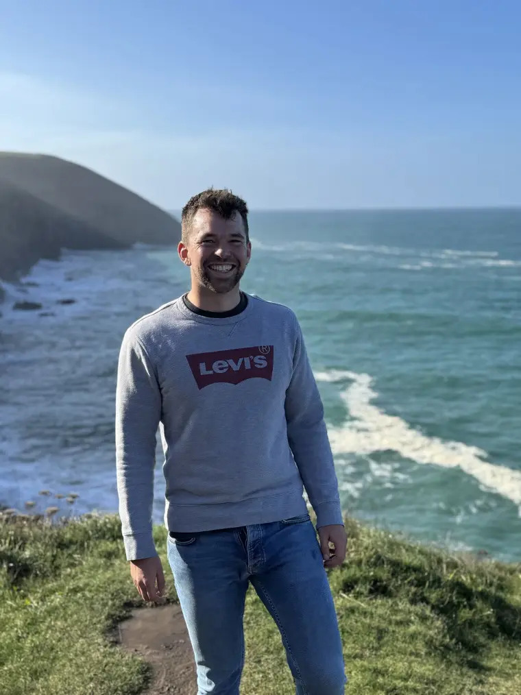 Joe Stone smiling on a clifftop by the sea.